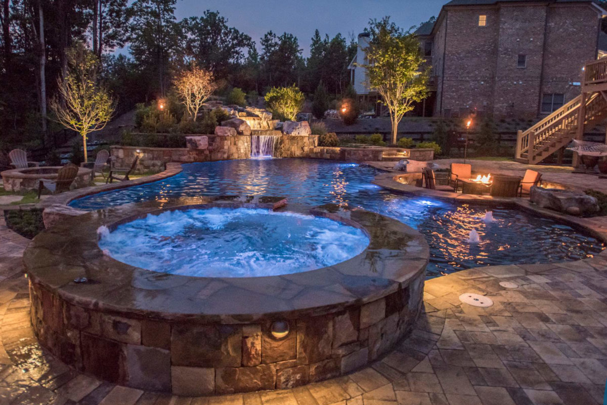 photo of a backyard round pool and jacuzzi design with a waterfall feature in PebbleTec Black Pearl Pool Finish dark blue water color taken in the evening
