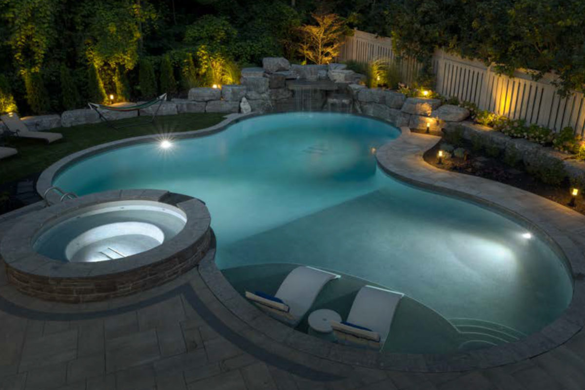 photo of a round pool with a rock waterfall feature in the evening in PebbleTec Moonlight Grey Pool Finish light blue water color for World's Greatest Pools 2018